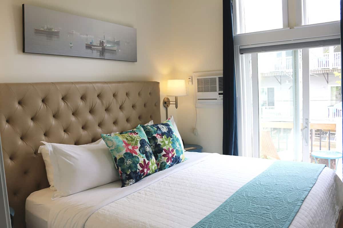 Quarto de hotel com cabeceira estofada, cama grande com almofadas brancas e florais, manta azul-esverdeada, janela com vista para edifícios e ar-condicionado fixado na parede.