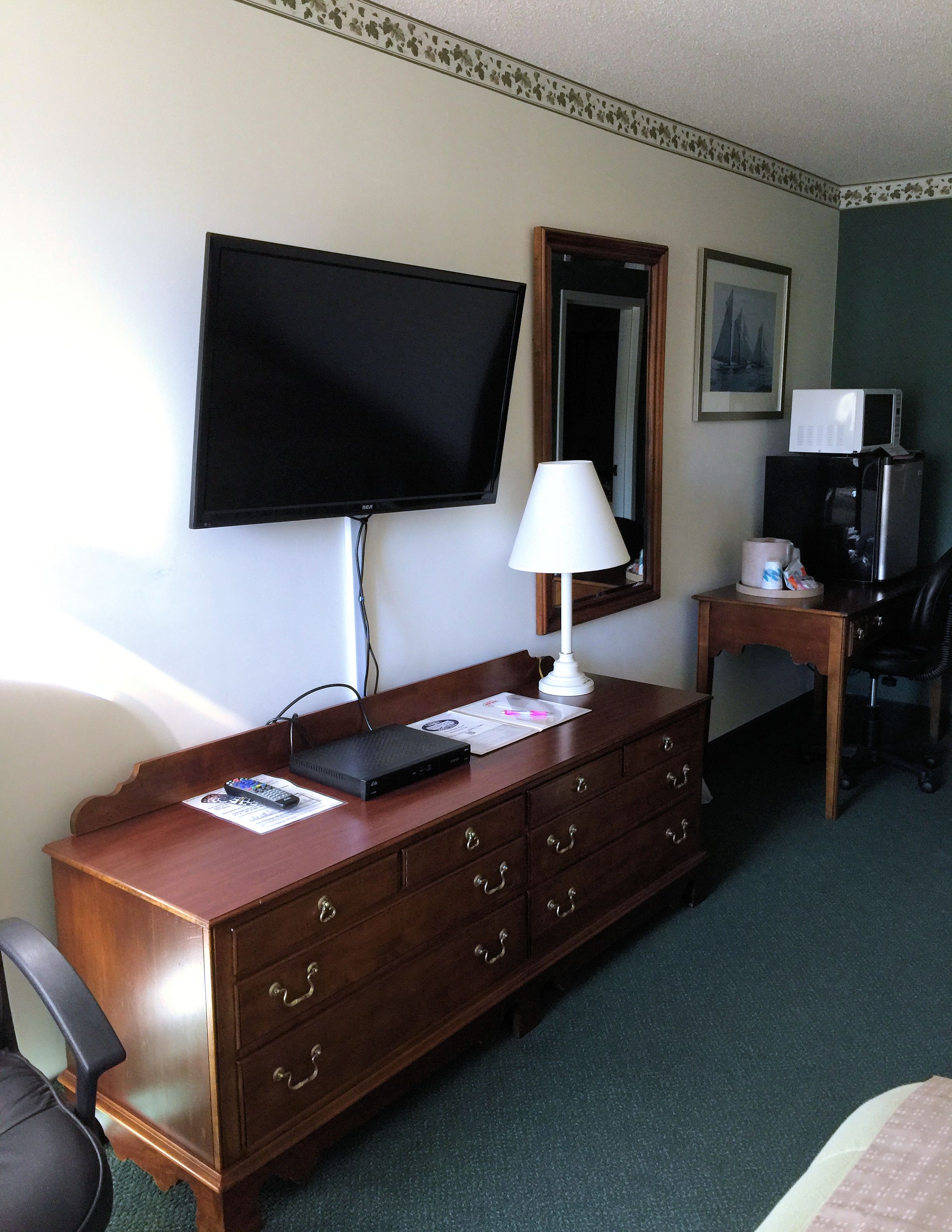 Une chambre d’hôtel avec une commode en bois, un téléviseur à écran plat fixé au mur, un bureau avec un four à micro-ondes, une chaise et un lit.