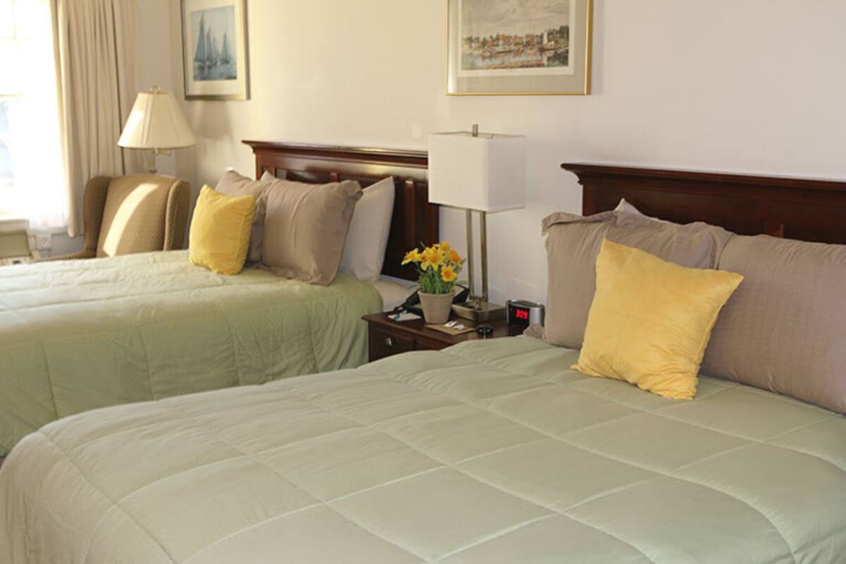 Duas camas com roupa de cama bege e amarela, uma mesa de cabeceira com abajur e um relógio.