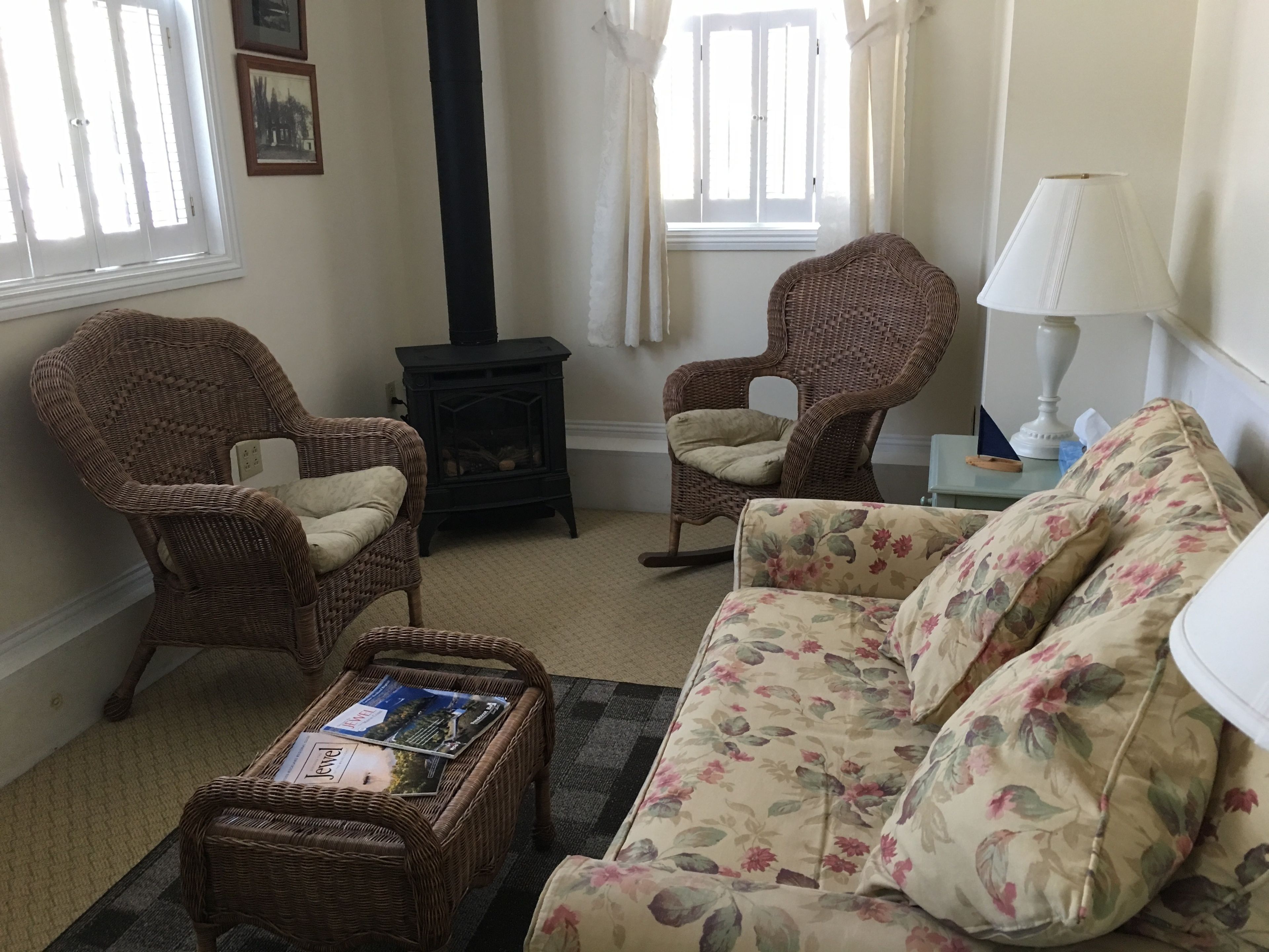 A cozy living room with wicker furniture, a floral sofa, a small table with magazines, and a black wood-burning stove.