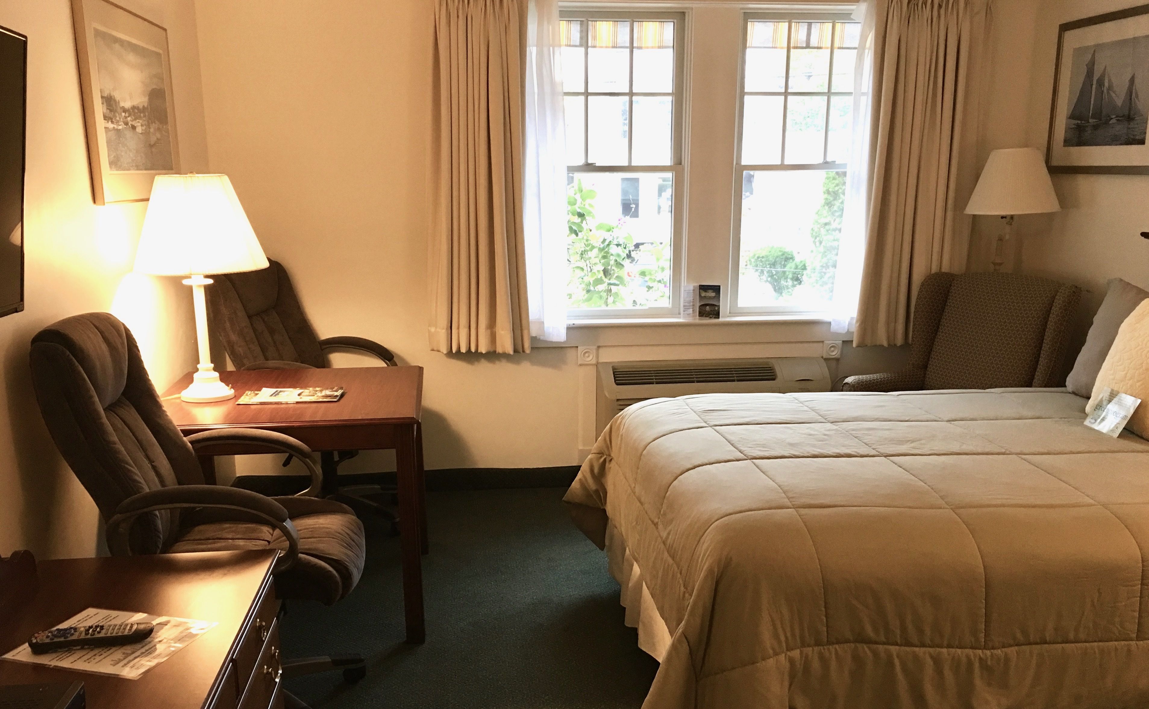 Une chambre d’hôtel avec un lit, un bureau avec une lampe, une chaise et une fenêtre avec des rideaux.