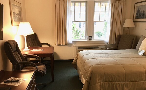 A hotel room with a bed, a desk with a lamp, a chair, and a window with curtains.