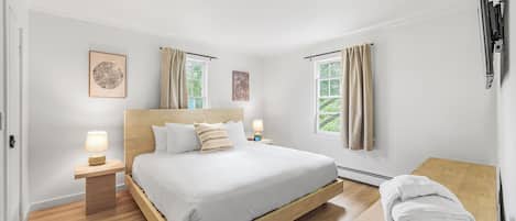 Una habitación de hotel moderna con una cama de madera, dos mesitas de noche con lámparas, vistas a zonas verdes a través de las ventanas y una toalla doblada con cuidado sobre un banco de madera.