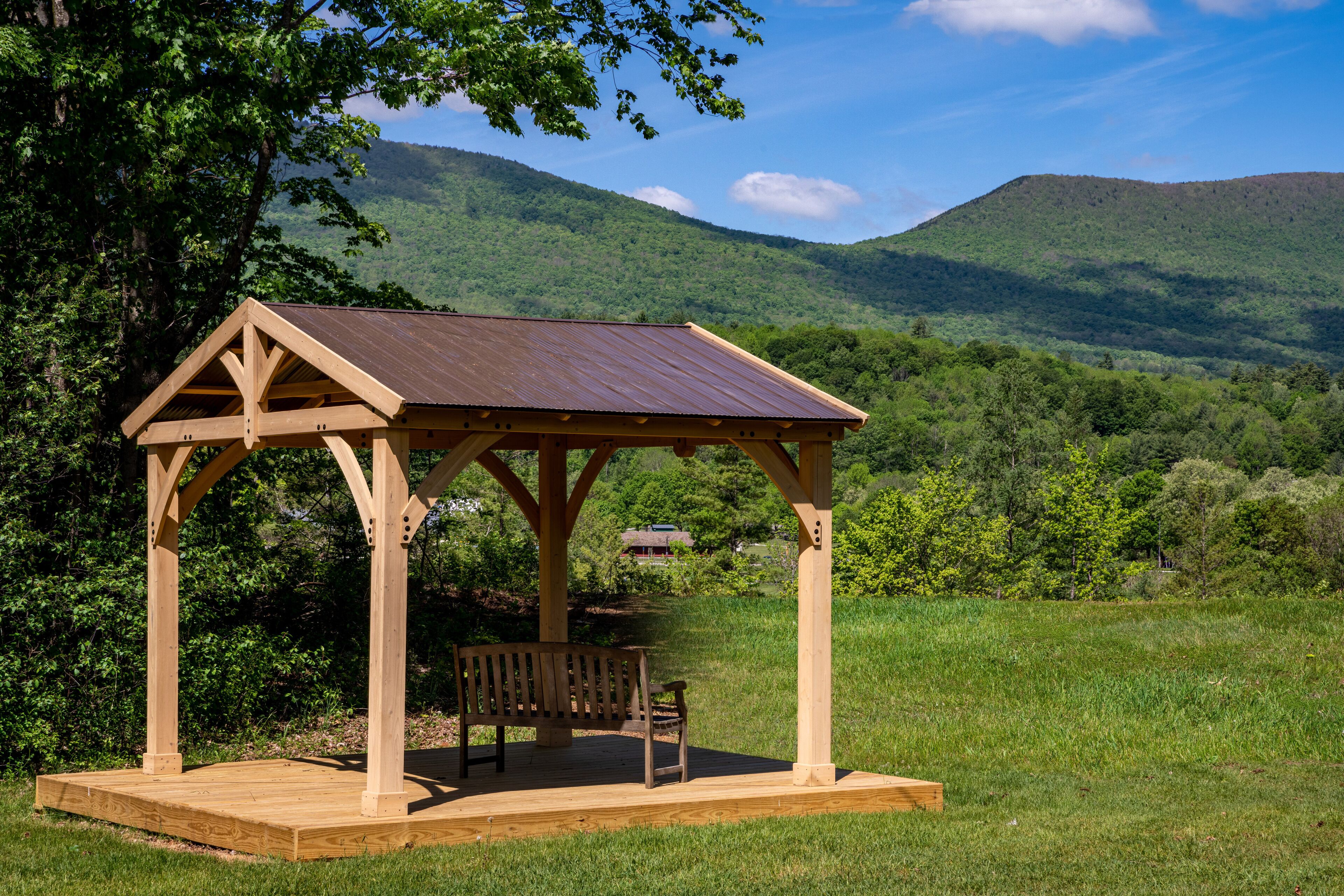 A wooden gazebo with a bench, set in a grassy field with a mountain backdrop.