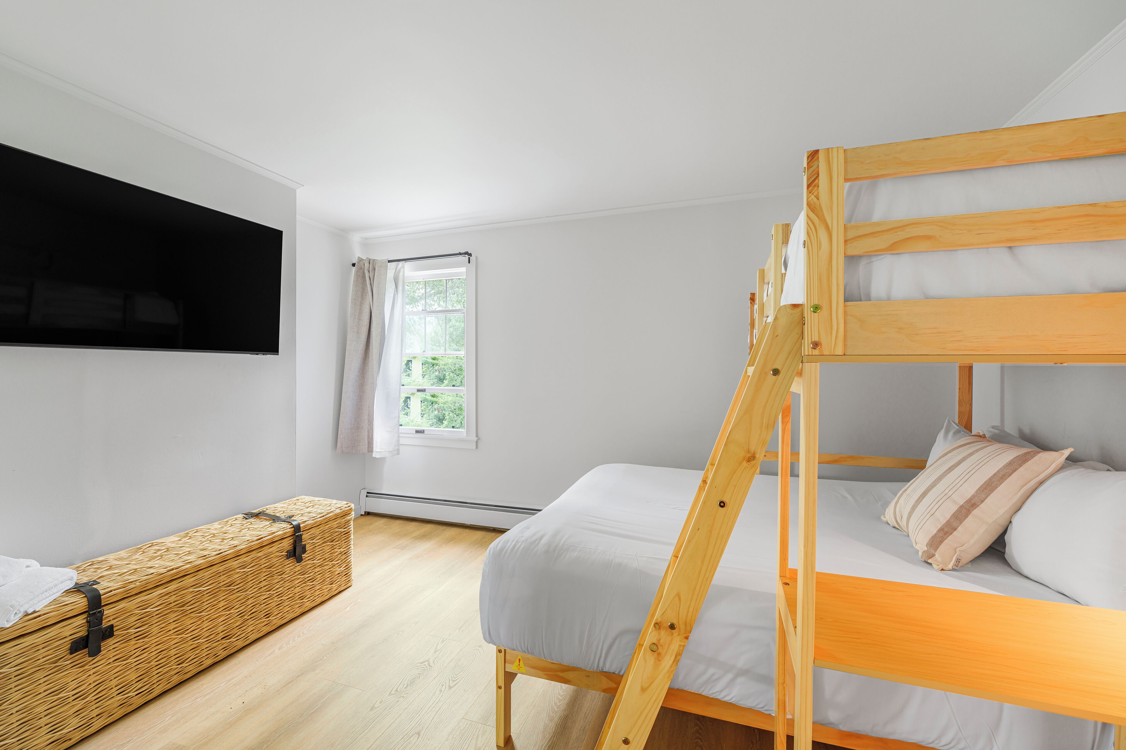 Une chambre avec un lit superposé, un téléviseur fixé au mur, une fenêtre avec des rideaux et une commode en bois.