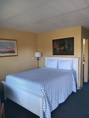 A hotel room with a bed, a painting, a lamp, and a door.