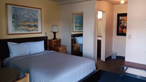 A hotel room with a bed, bedside table, lamp, artwork on the walls, and a bathroom visible through an open door.