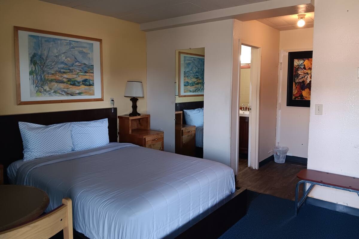 Habitación de hotel con cama, mesita de noche, lámpara, obras de arte en las paredes y un baño visible a través de una puerta abierta.