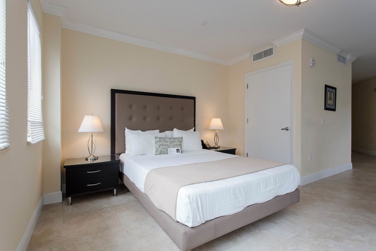 A large bed with a tufted headboard, two bedside tables with lamps, a door, and a framed picture on the wall.