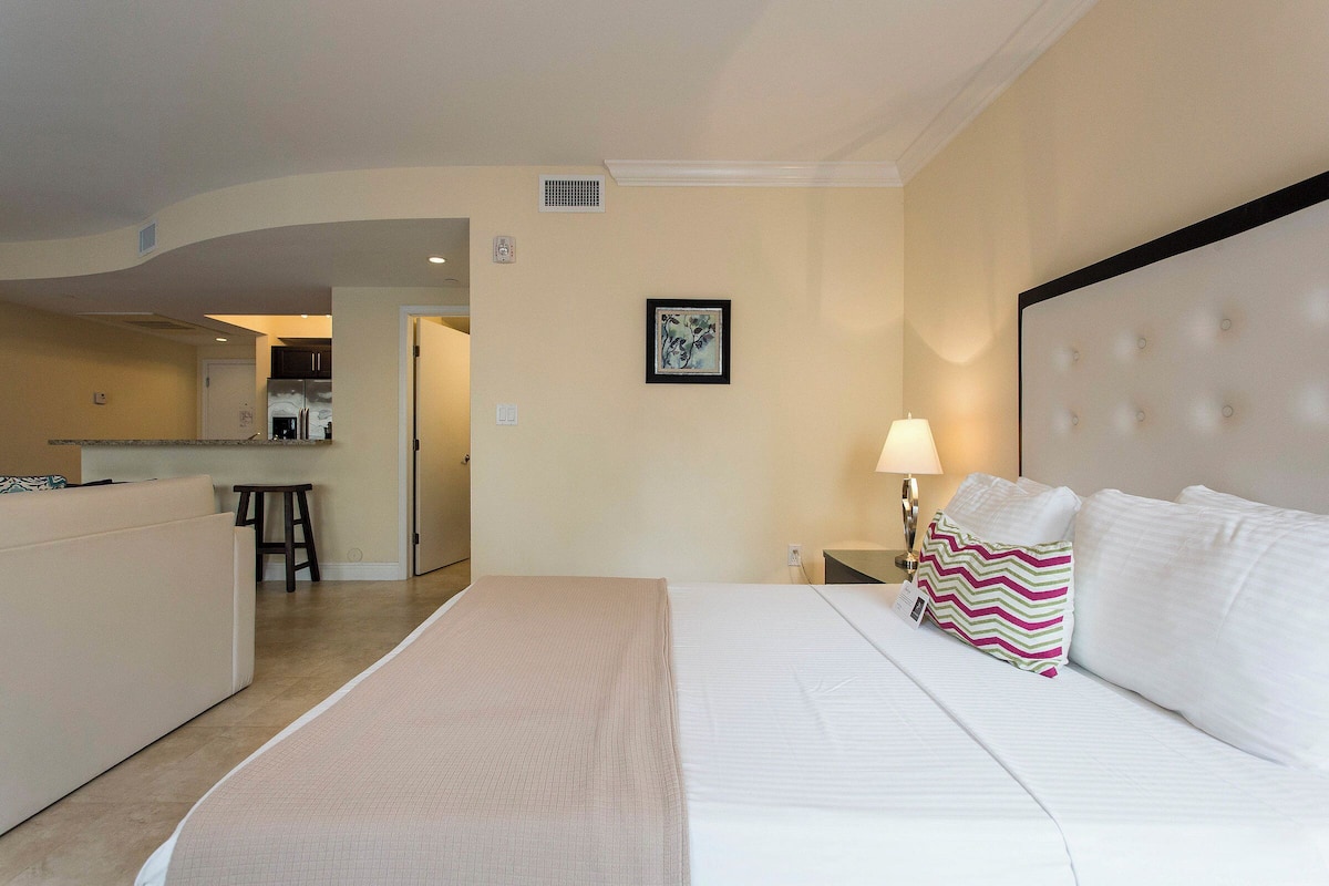A hotel room with a bed, a nightstand, a lamp, a picture on the wall, and a kitchenette in the background.