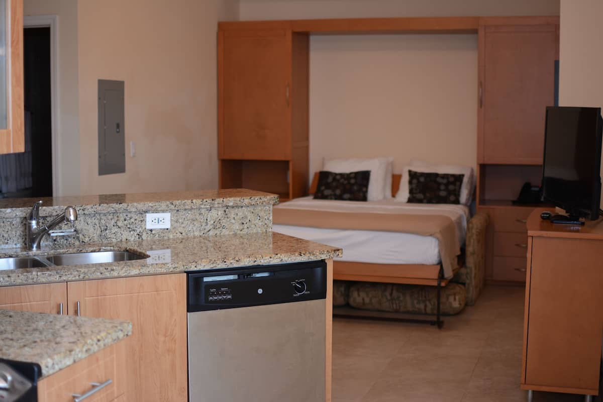 A compact hotel room with a kitchenette, a bed with two pillows, a television, and a small cabinet.