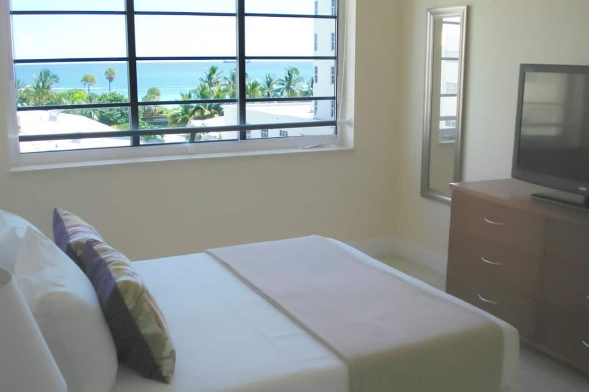A hotel room with a bed, a television on a wooden cabinet, and a window with a view of palm trees and the ocean.