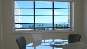 A dining area with a round glass table and four chairs, a window with a view of the ocean and palm trees, and a building in the background. - Westover Arms Hotel  (Miami Beach)
