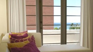 A modern living room with a white sofa, a purple patterned throw pillow, and a view of a beach and ocean through large windows. - Westover Arms Hotel  (Miami Beach)