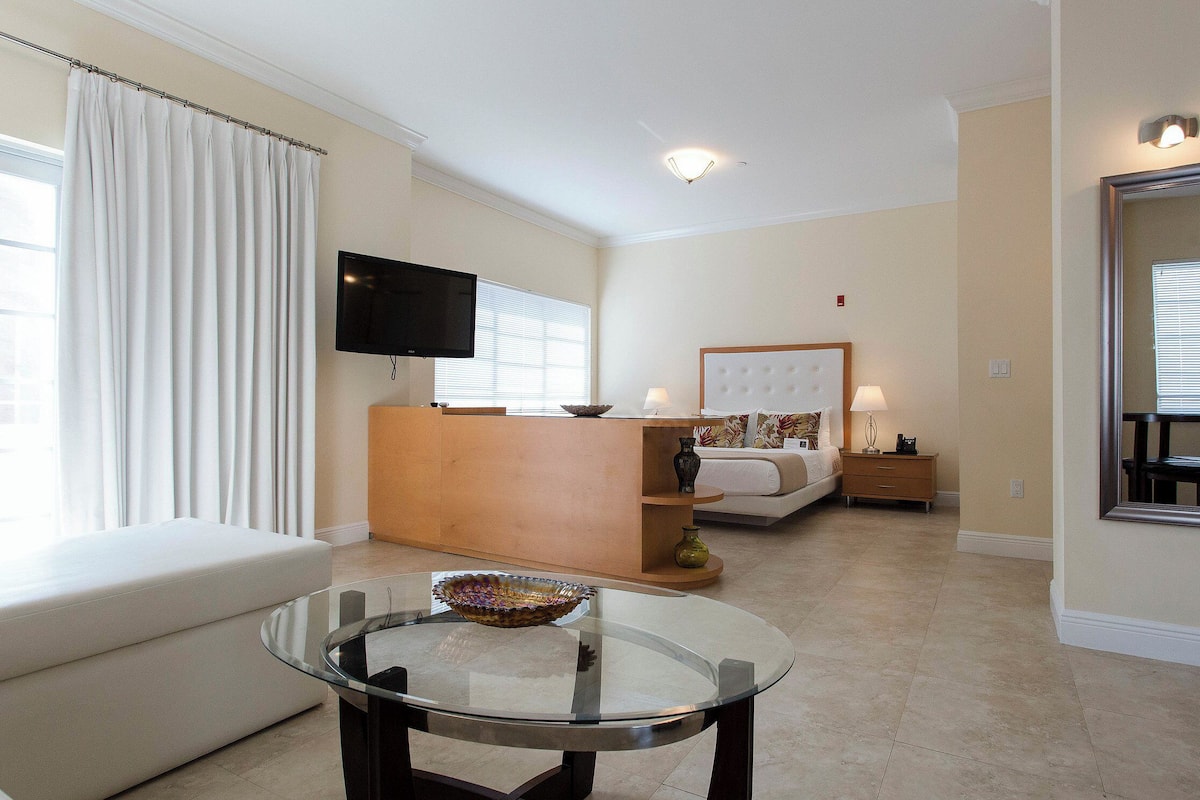 A hotel room with a glass table, a sofa, a bed with a headboard, a television, and a mirror.