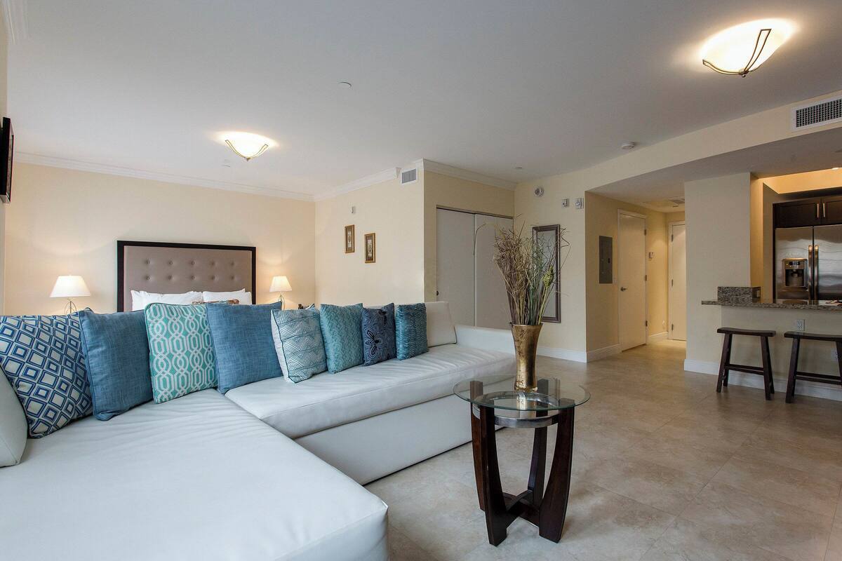 A modern living room with a sectional sofa, a glass coffee table, and a kitchen area in the background.