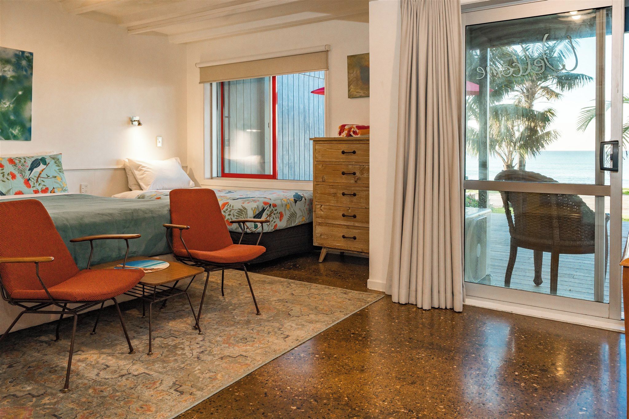 A room with a bed, two chairs, a dresser, and a view of the ocean through a glass door.