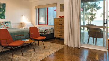 A room with a bed, two chairs, a dresser, and a view of the ocean through a glass door.