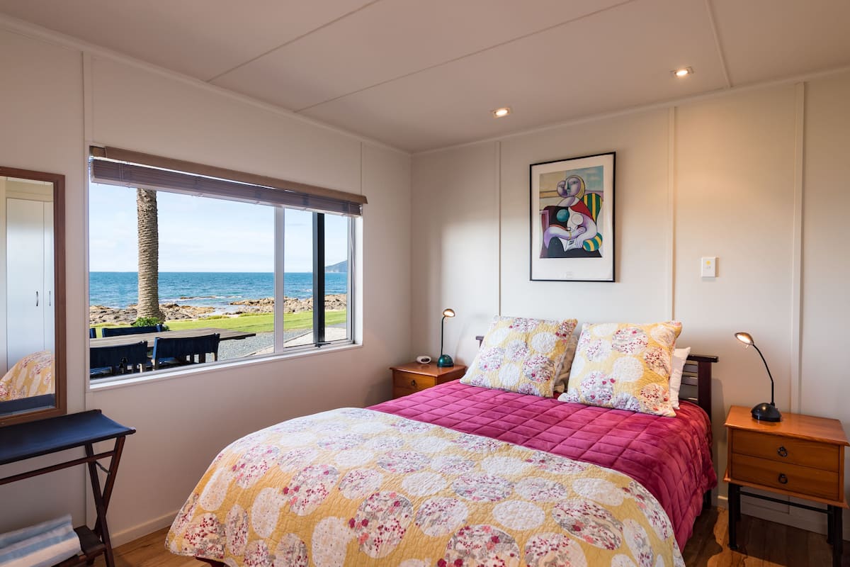 A bedroom with a large bed, a window with a view of the beach, a desk, and a nightstand.