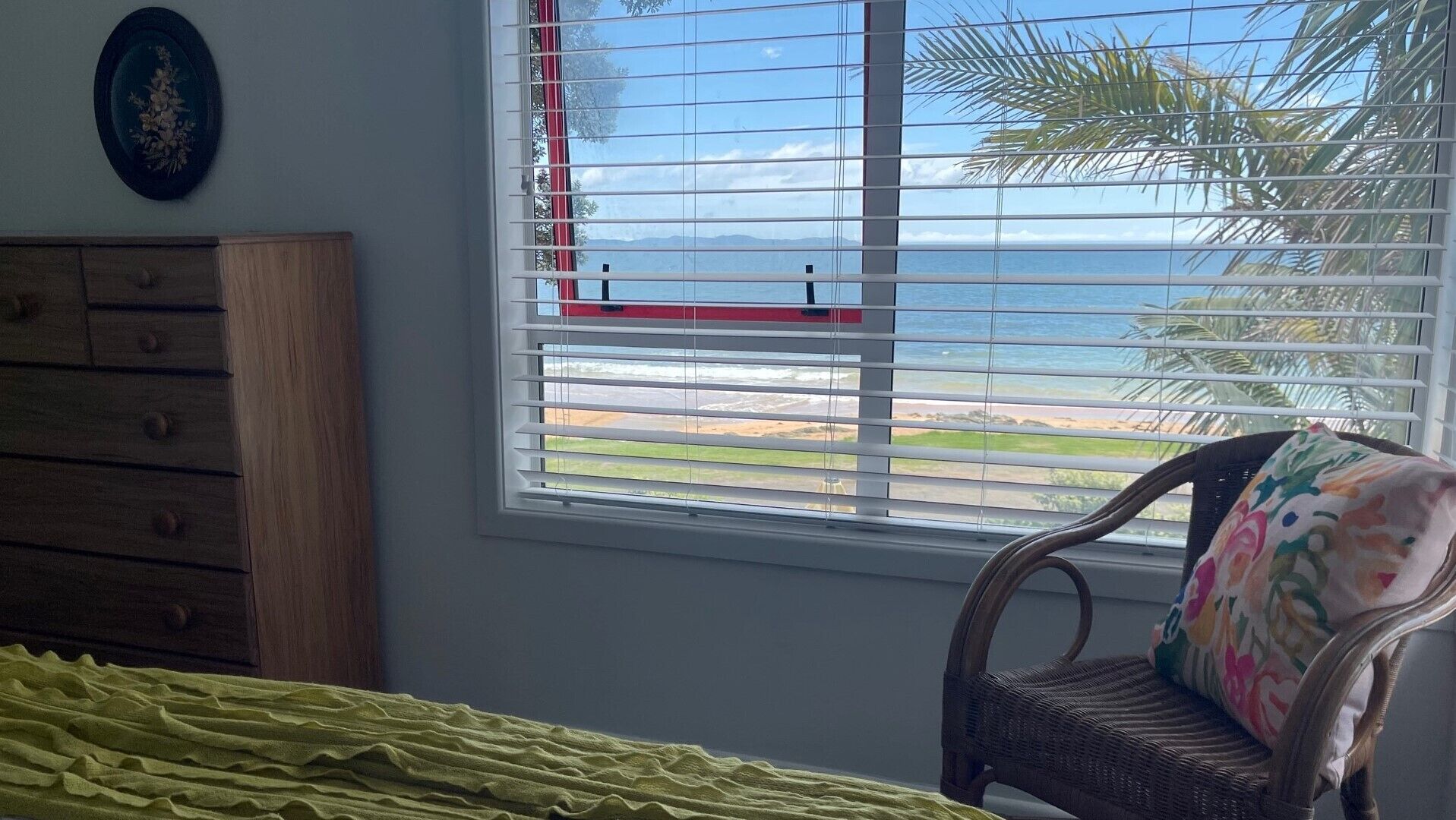 A room with a bed, a dresser, a chair with a colorful cushion, and a window with a view of the ocean and palm trees.