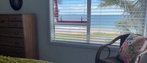 A room with a bed, a dresser, a chair with a colorful cushion, and a window with a view of the ocean and palm trees.
