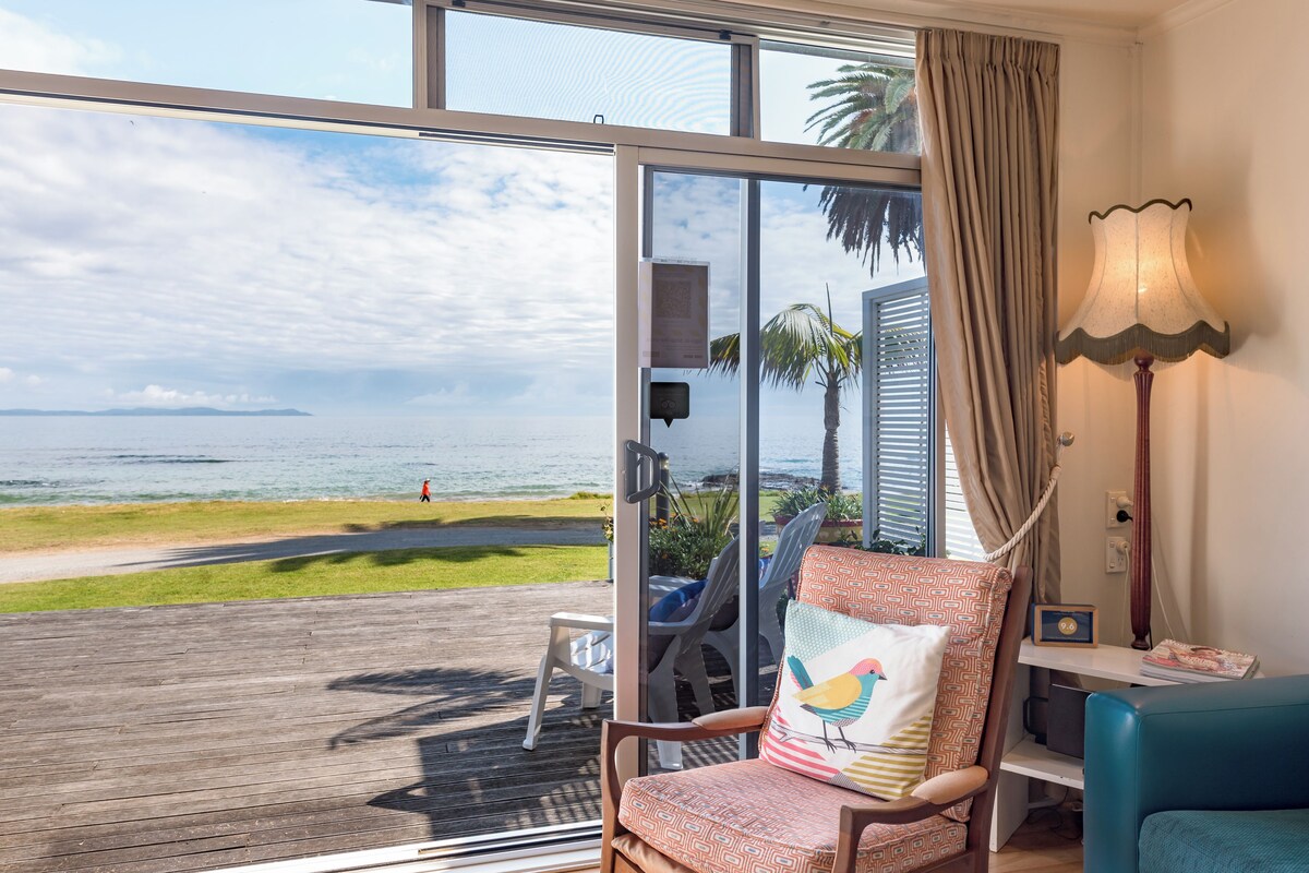 A room with a view of the beach, a chair with a patterned cushion, a lamp, and a small table.