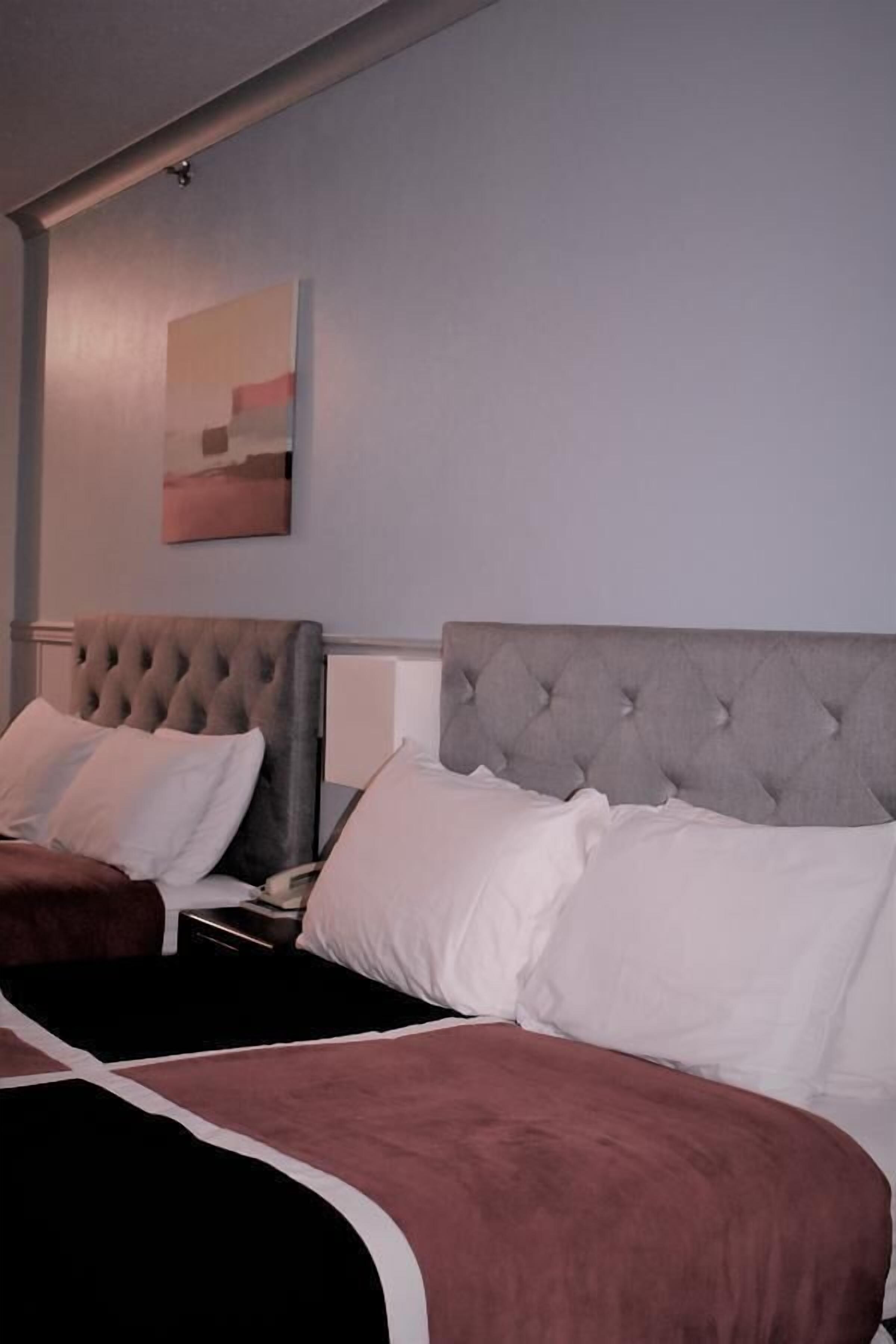 Habitación de hotel con dos camas, cada una con cabecero capitonado, ropa de cama blanca y marrón, y una mesita de noche con lámpara.
