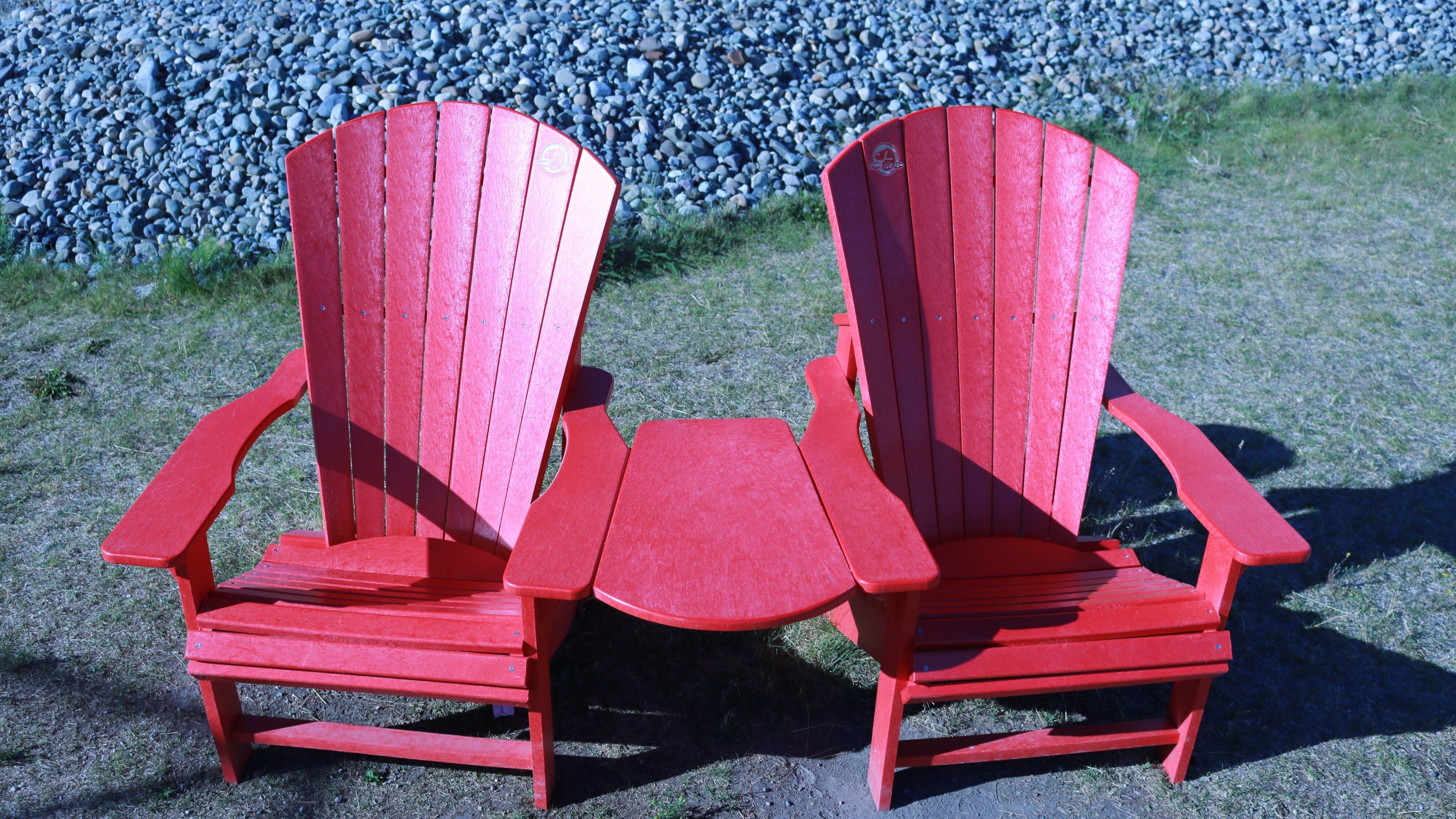 Dos sillas Adirondack rojas con una pequeña mesa entre ellas, sobre un suelo de grava con césped al fondo.