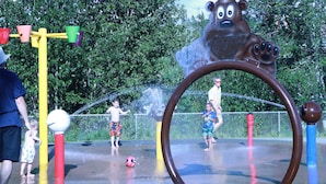 A water play area with a large bear sculpture, a water spout, and a metal fence.