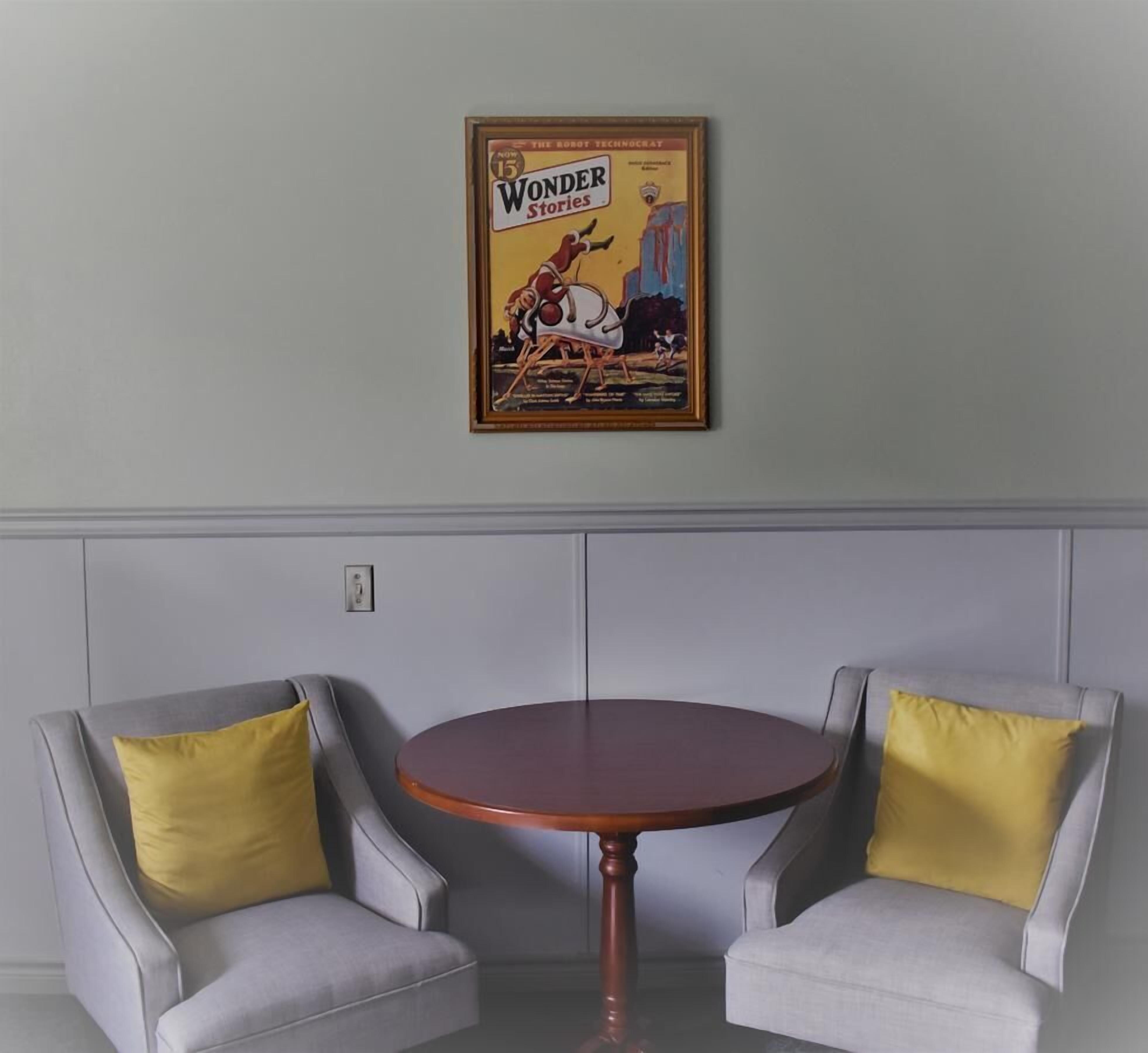 Ein Zimmer mit zwei grauen Sesseln, einem runden Holztisch und einem gerahmten Poster an der Wand.