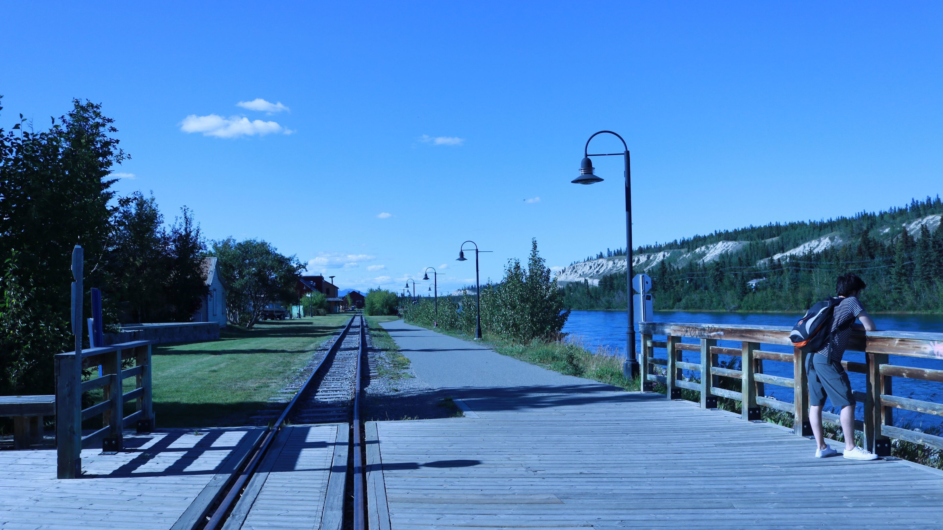A wooden boardwalk with a railway track, a person leaning on a railing, and a lake with a forested hillside in the background.