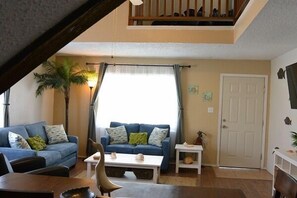 A living room with two blue sofas, a coffee table, and a dining table. There is a staircase leading to an upper level. - Gulf 4 Bedroom Condo by Fountain Vista Homes (South Padre Island)
