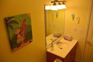 A bathroom with a yellow wall, a painting of a tropical scene, a sink with a mirror, and a towel rack.