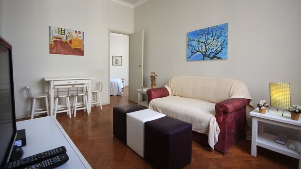 Een woonkamer met een bank, poefs, een televisie, een eettafel met stoelen, en kunst aan de muren.