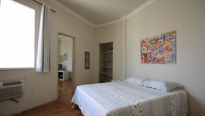 Een slaapkamer met een bed, een radiator, een raam met gordijnen, een deur die naar een andere kamer leidt, en een kleurrijk abstract schilderij aan de muur.