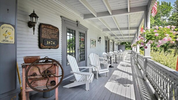 A covered porch with white Adirondack chairs, a vintage washing machine, and a plaque from 1874. - Auberge Lakeview Inn (Lac-Brome)