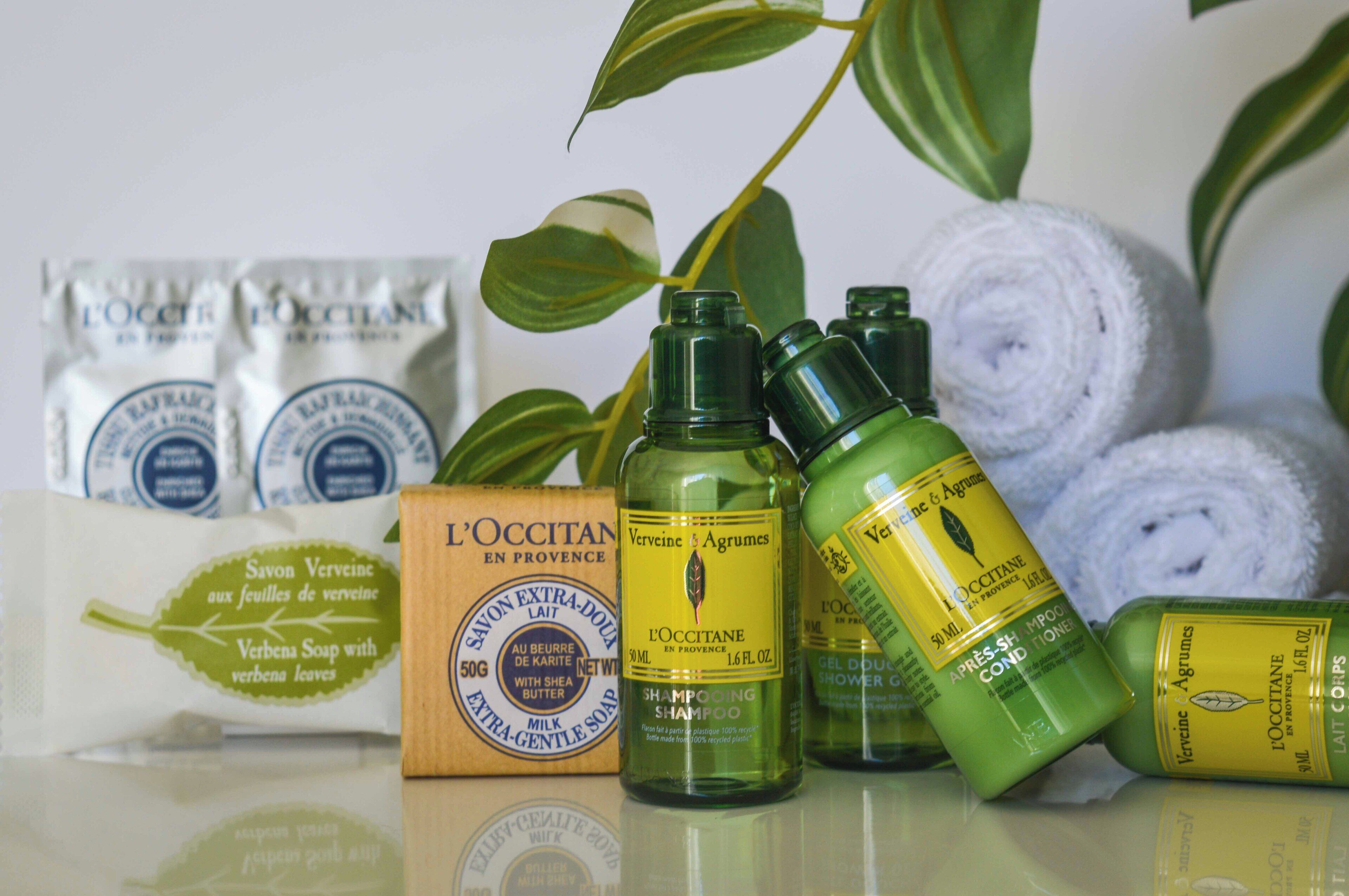 Des articles de toilette, notamment du shampoing, de l’après-shampoing et du savon, sont disposés sur une surface réfléchissante, avec une plante et des serviettes enroulées en arrière-plan.