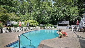 A swimming pool with a tiled edge, surrounded by white chairs and umbrellas, with a wooden deck and a white fence in the background. - Auberge Lakeview Inn (Lac-Brome)