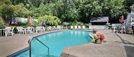 Piscina com borda azulejada, rodeada por cadeiras e guarda-sóis brancos, com um deck de madeira e uma cerca branca ao fundo.