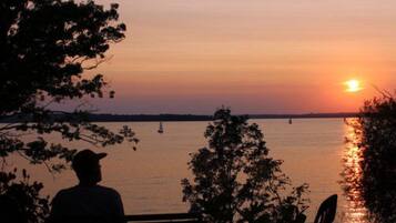 A sunset view from a dock with a person sitting on a bench, a sailboat in the distance, and a calm body of water.