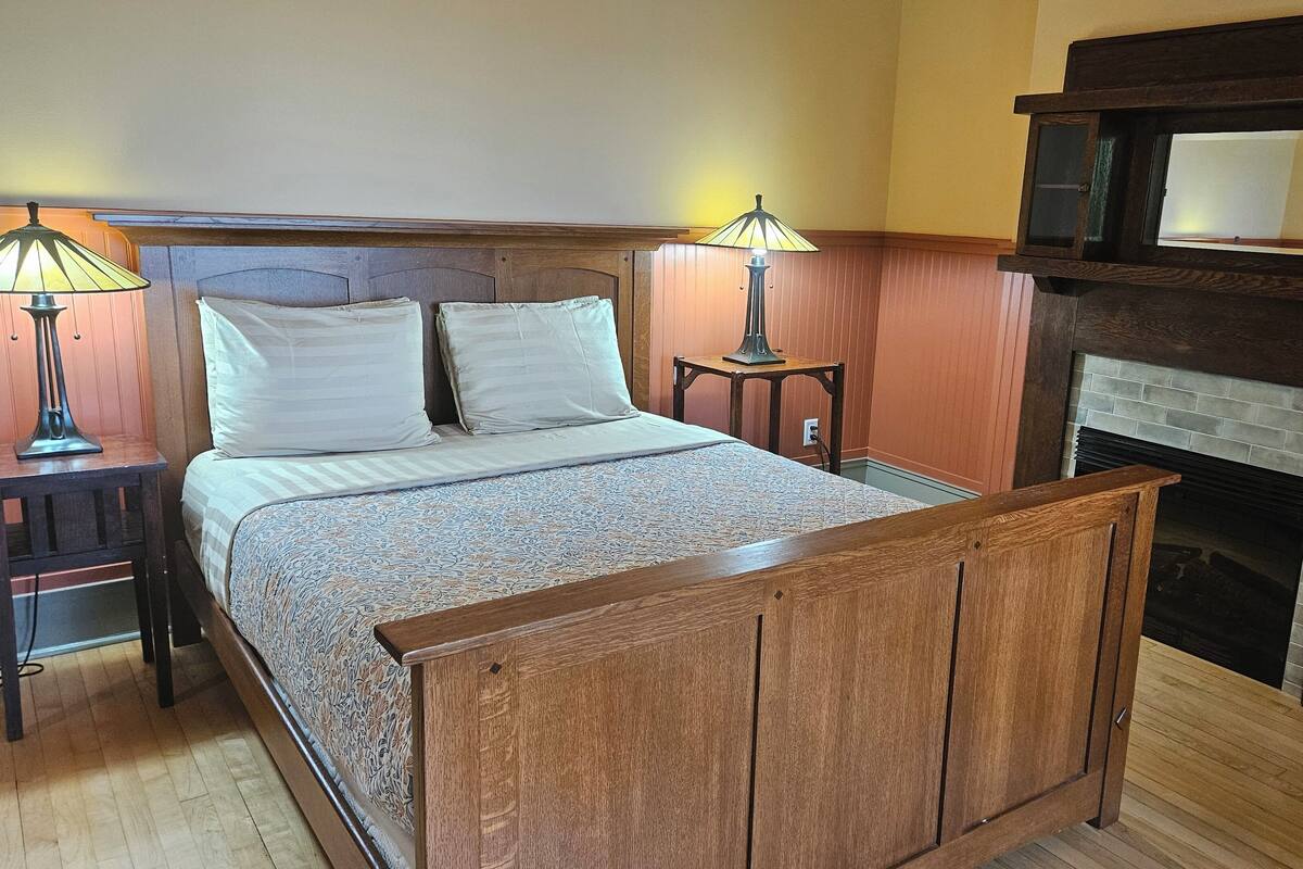 A bedroom with a wooden bed, two bedside lamps, a fireplace, and a nightstand.