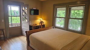 A bedroom with a bed, a desk with a lamp, a TV, and large windows with a view of greenery.