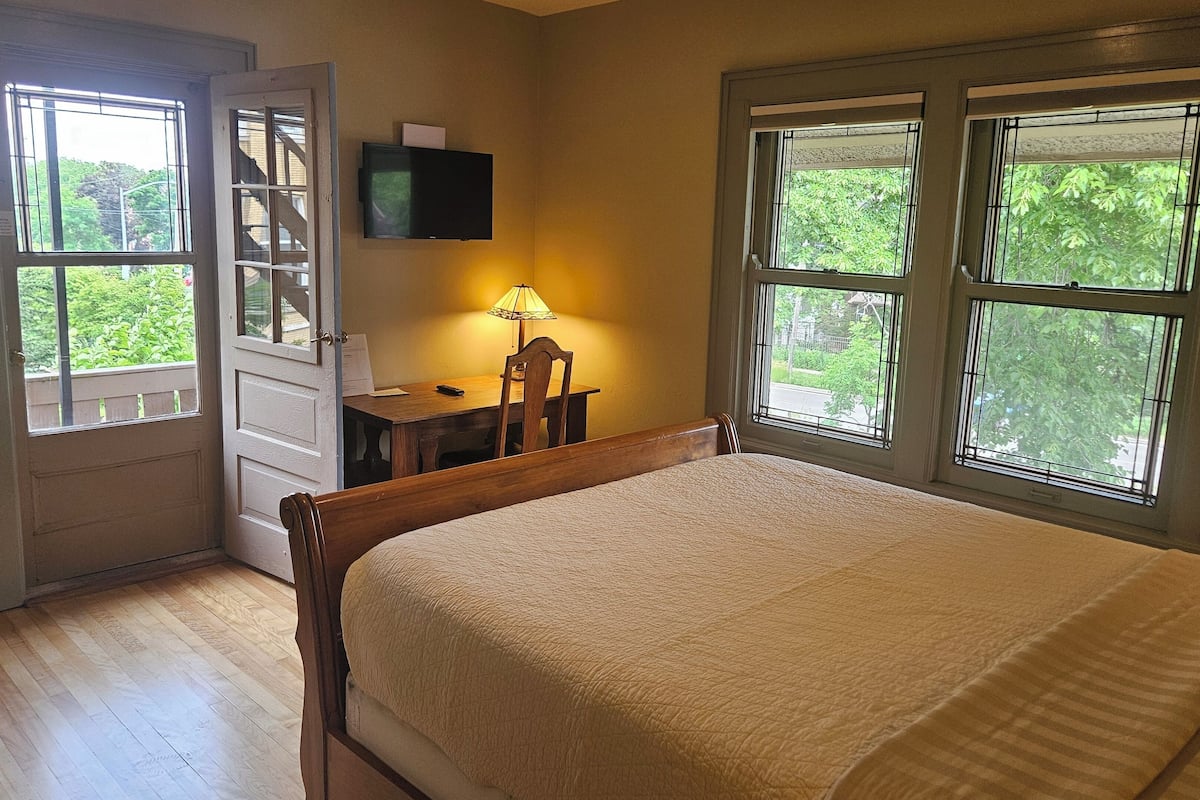 A bedroom with a bed, a desk with a lamp, a TV, and large windows with a view of greenery.