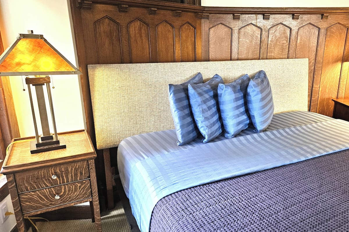 A neatly made bed with a striped blue bedspread and four pillows, a wooden headboard, a nightstand with a lamp, and a wooden dresser.