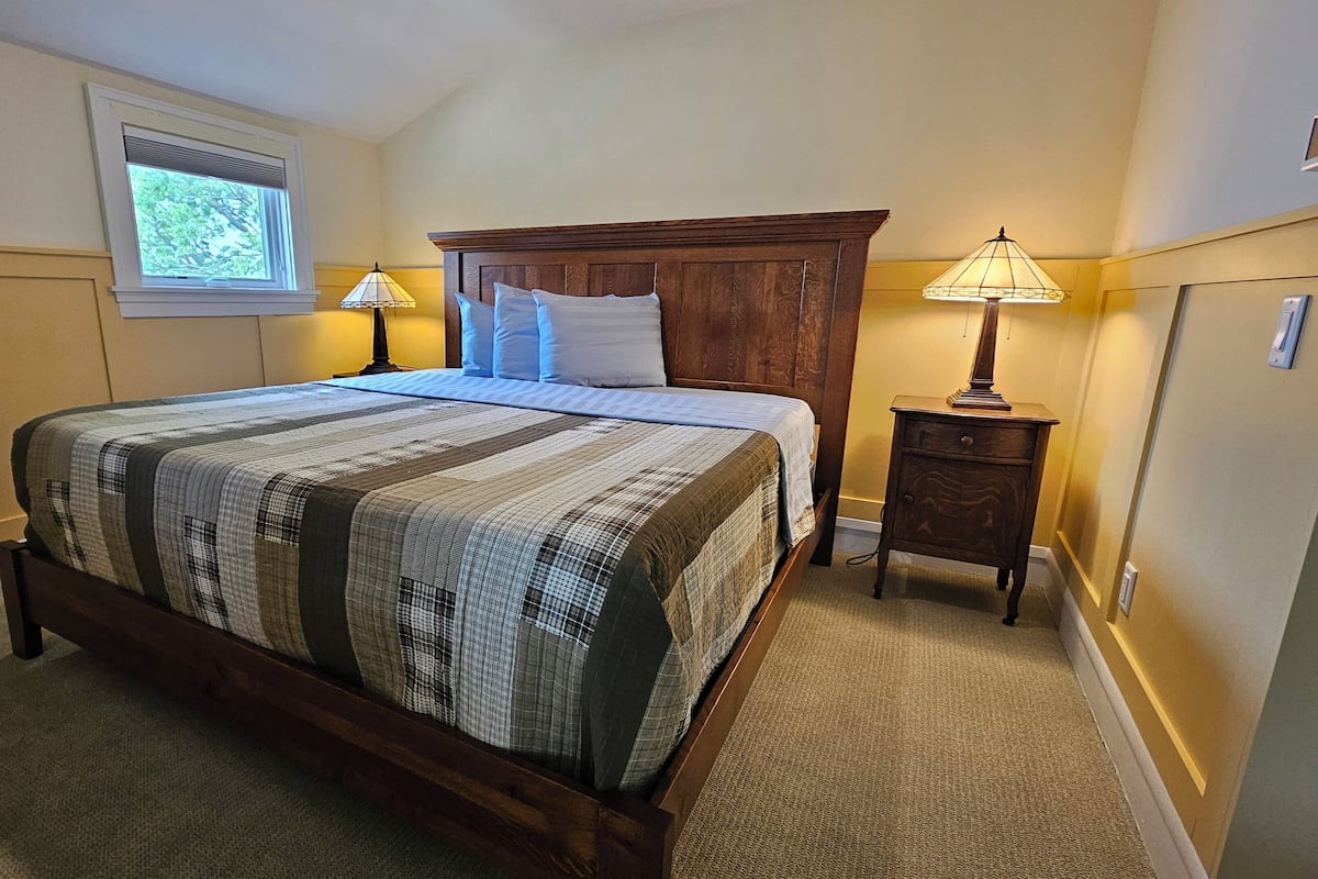 A bedroom with a large bed, wooden headboard, two bedside lamps, a window with blinds, and a carpeted floor.