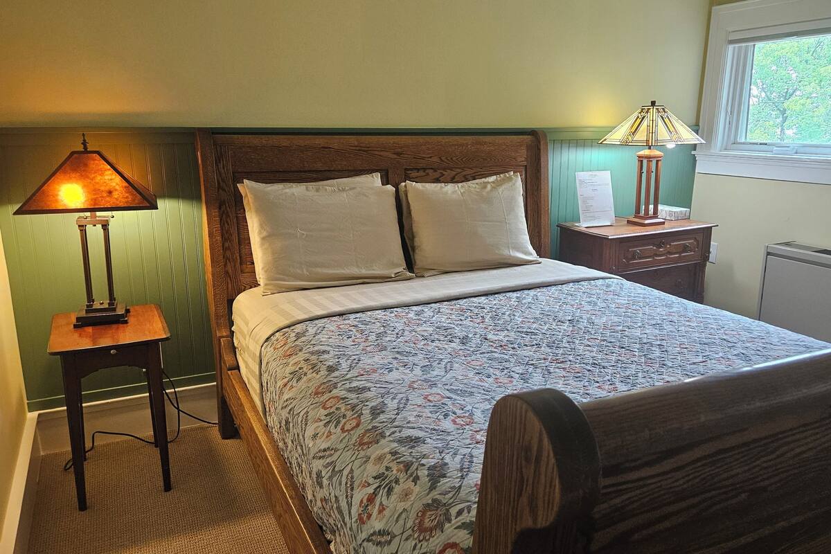 A bedroom with a wooden bed, two bedside lamps, a window with a view of trees, and a radiator.