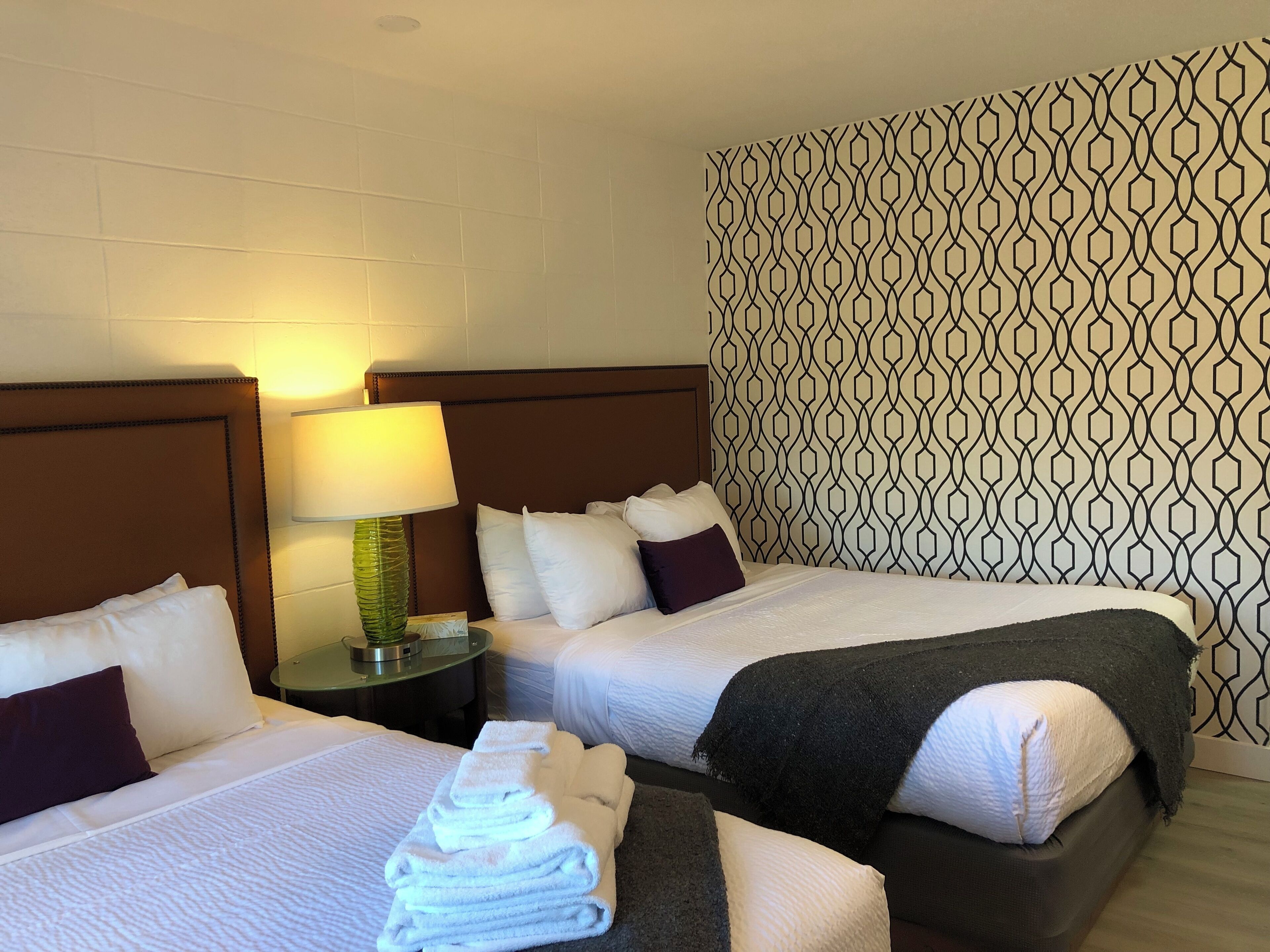 Habitación de hotel con dos camas, una mesita de noche con lámpara y papel tapiz estampado.