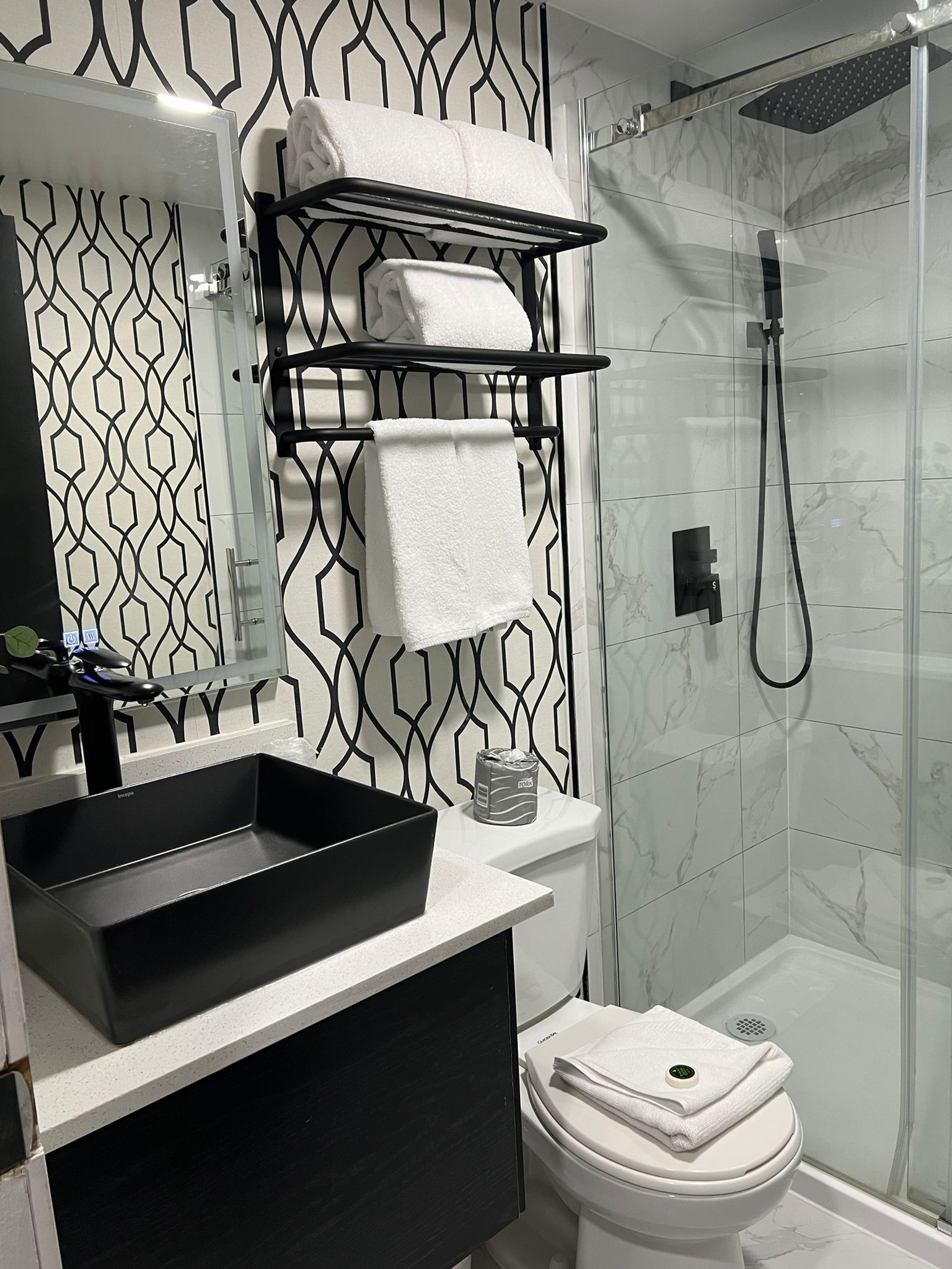 A modern bathroom with a black and white patterned wall, a black sink, a toilet, and a glass-enclosed shower area.