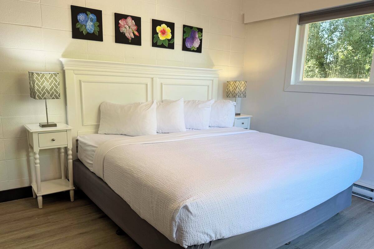 Una cama bien hecha con sábanas blancas, un cabecero blanco y dos mesitas de noche con lámparas. Cuatro cuadros florales adornan la pared sobre la cama.