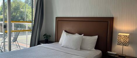 A hotel room with a large bed, a nightstand with a lamp, a potted plant, and a view of the outdoors through a sliding glass door.